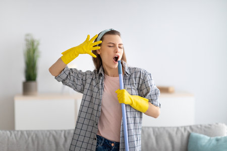 Carefree young housewife in rubber gloves using mop as mic and singing, cleaning modern apartment. Millennial maid having fun during domestic chores, enjoying her favorite songの写真素材