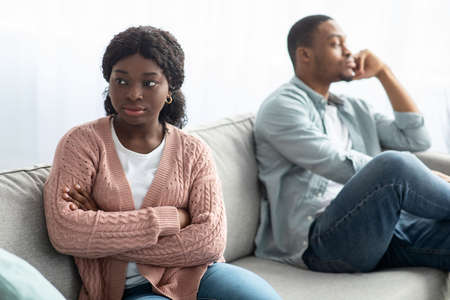 Angry black woman sitting apart from her frustrated boyfriend, young african american couple having fight at home, suffering from difficulties in relationships during  self-isolationの写真素材