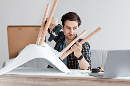 Instructions and online blog about home renovation and furniture assembly. Serious handsome young male screwing bolts in new chair with electric screwdriver, looking at laptop in living room interiorの写真素材