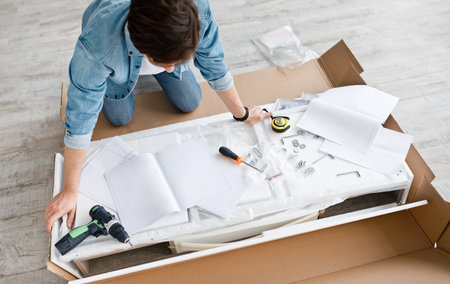 Assembly of new home furniture, construction equipment and repairs. Busy young guy reads instructions on floor in living room interior, near details, tools in daylight, top view, empty space, croppedの写真素材