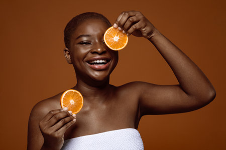 Portrait Of Cheerful Black Lady Wrapped In Towel Posing With Orange Fruit Slices Near Face, Attractive African American Female Enjoying Vitamic C For Healthy Skin, Standing Over Brown Backgroundの写真素材