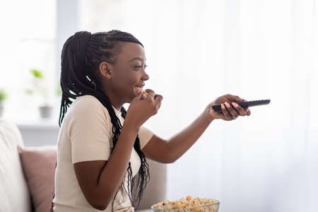 Side view of joyful african american woman watching television and eating popcorn at home, closeup,...