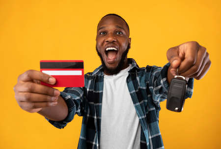 Make Dream Come True. African Man Showing New Car Key And Credit Card Buying Automobile Standing Over Yellow Studio Background. Auto Loan, Card-Based Payment Transaction And Banking Serviceの写真素材