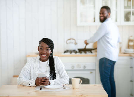 Young Black Woman Sitting At Table In Kitchen With Smartphone While Her Husband Cooking Lunch On Background, Millennial African American Couple Spending Time Together At Home, Selective Focusの写真素材