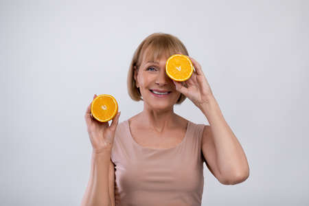Natural fruit vitamins for mature skin. Smiling senior woman holding fresh orange in front of her eye over light studio background. Beauty and organic cosmetics conceptの写真素材