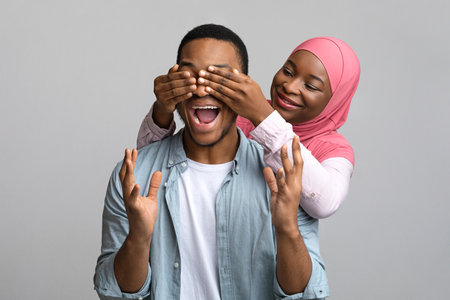 Pretty black lady in hijab covering her emotional boyfriend eyes over grey studio background, copy space. Cheerful african american muslim woman making surprise for her beloved manの写真素材