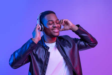 Music Lover. Handsome Black Guy In Wireless Headphones And Leather Jacket Listening His Favorite Songs, Relaxed African American Man Standing With Closed Eyes Under Neon Light Over Purple Backgroundの写真素材