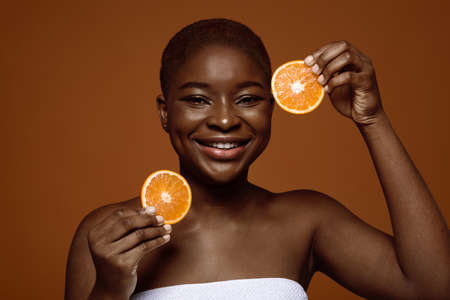 Vitamin C For Skin. Attractive Black Lady Wrapped In Towel Holding Orange Fruit Slices Near Face, Smiling African Woman Enjoying Organic Components For Cosmetics, Standing On Brown Background, Closeupの写真素材