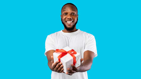 African Man Offering Wrapped Gift Box Smiling To Camera Posing Over Blue Studio Background. Birthday Gifts And Presents, Holiday Celebration Concept. Panoramaの写真素材