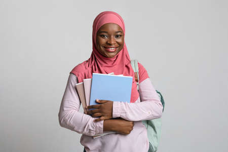Pretty african american female student young woman in hijab posing with notebooks and books over grey studio background, carrying backpack, cheerfully smiling at camera, copy spaceの写真素材