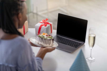 Closeup of unrecognizable african american woman holding birthday cake, using laptop with blank screen, sitting at desk with gift box and glass of champagne on it, home interior, over shoulder shotの写真素材
