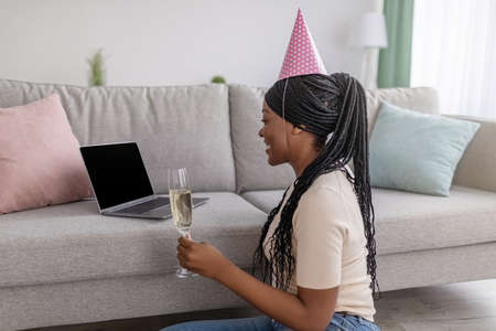 Young african american lady having online birthday party from home during coronavirus pandemic, wearing party hat, holding glass of champagne and looking at empty laptop screen, copy spaceの写真素材