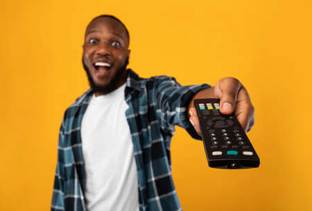 Excited African American Man Pointing Television Controller To Camera Switching Channels Watching TV Standing Over Yellow Studio Background. Television Programming Advertisement. Selective Focusの写真素材