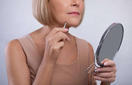 Cropped view of mature woman removing unwanted hair from her chin, using tweezers, looking in mirror on light studio background. Unrecognizable senior lady taking care of her appearanceの写真素材