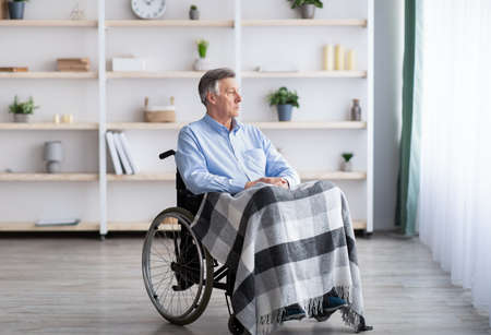 Sad disabled senior man sitting on wheelchair alone at retirement home, looking through window....