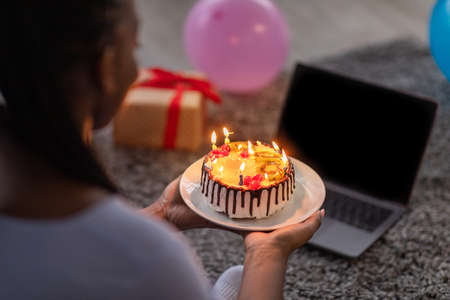 Online birthday party. Closeup of black woman holding birthday cake with lit candles, using laptop with empty screen, decorated with balloons and gift boxed home interior, over shoulder shotの写真素材