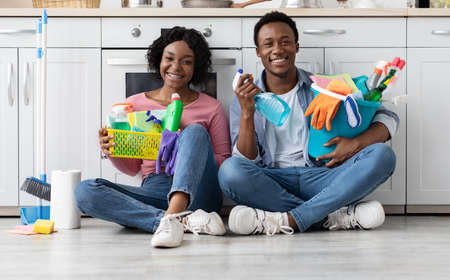 Smiling black couple sitting in kitchen, holding house-keeping toolsの写真素材