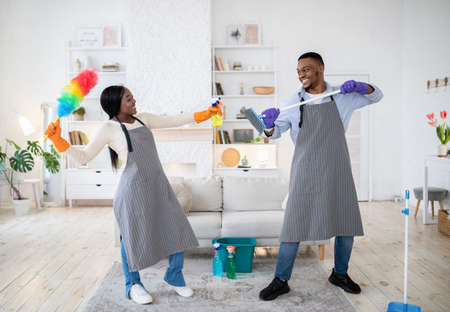 Playful black couple having mock fight with mop and detergent, being silly during house cleanup, indoorsの写真素材