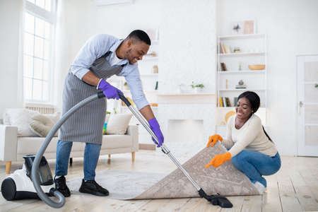 Positive black couple cleaning up together, vacuuming under carpet at their house, copy spaceの写真素材