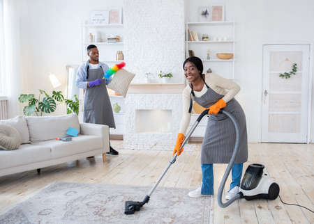 Happy black woman vacuuming floor while her boyfriend dusting cushion at their apartment, copy spaceの写真素材
