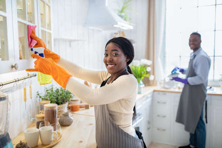 Happy black lady cleaning kitchen cabinet, her husband washing dishes on backgroundの写真素材
