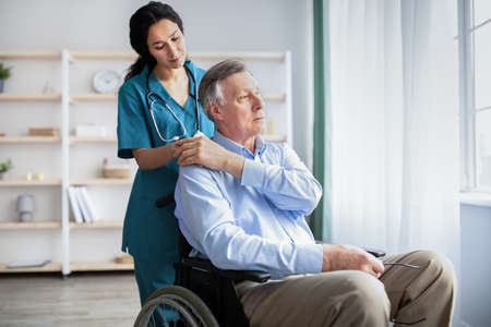 Young doctor holding elderly disabled males hand, giving him support at retirement home