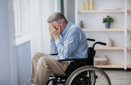 Depression and loneliness in disabled seniors. Elderly man in wheelchair crying near window indoorsの写真素材