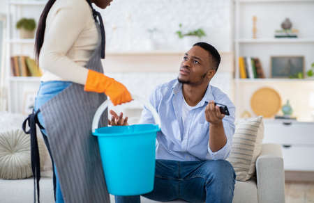 Lazy black man watching TV while his wife doing home cleanup, having fight with her, not wanting to help, indoorsの写真素材