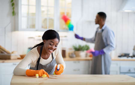 Joyful black woman cleaning kitchen table with detergent while her boyfriend dusting on background,...