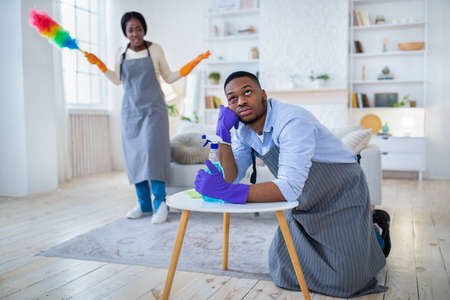 Young black guy feeling tired from housework, his angry wife scolding him for doing nothing, copy spaceの写真素材