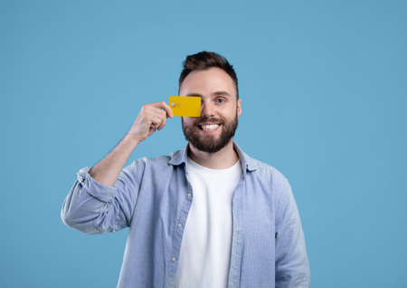 Happy young man in casual outfit holding credit card in front of his eye on blue studio background