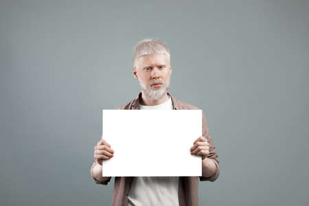 Albino man holding empty paper sheet with free space, posing to camera on grey background,...