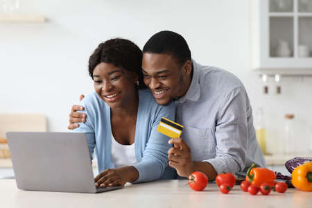 Joyful black couple using laptop and credit card in kitchenの写真素材