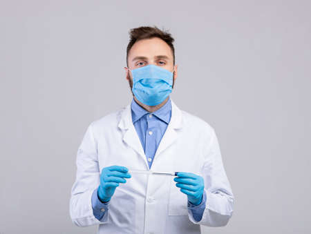 Young doctor wearing medical face mask and rubber gloves, holding coronavirus PCR test tube on grey studio backgroundの写真素材