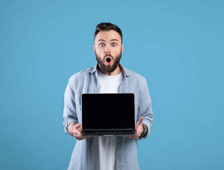 Shocked bearded guy showing laptop with blank screen on blue studio background, mockup for your websiteの写真素材
