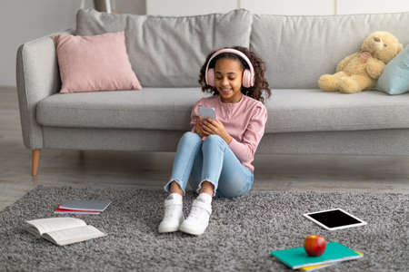 Happy african american girl listening to music in wireless headphones and chatting on her smartphoneの写真素材