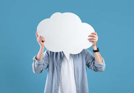 Faceless young man covering his face with speech bubble on blue studio background, mockup for designの写真素材