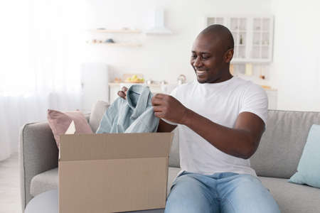 Shopping delivery. Mature african american man receiving clothes, unpacking cardboard box and holding new t-shirtの写真素材