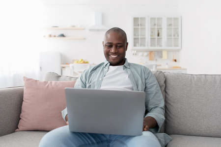 Free time. Happy middle-aged black man using laptop while resting on sofa at homeの写真素材