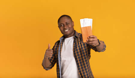 Border opening journey with air travel after covid-19. Smiling middle aged african american male showing thumb up and holding passport with air tickets, isolated on orange background, studio shotの写真素材