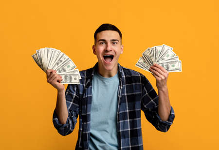 Cool hipster guy holding two fans of money, screaming WOW in excitement on orange studio backgroundの写真素材