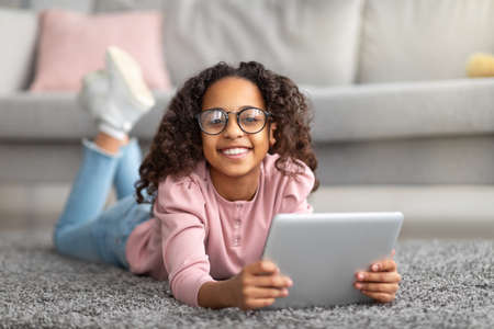 Cheerful homelanders girl using digital tablet, lying on carpet and smiling to camera