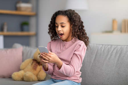 Surprised black girl receiving good news online in smartphone, resting at home and lying on carpet