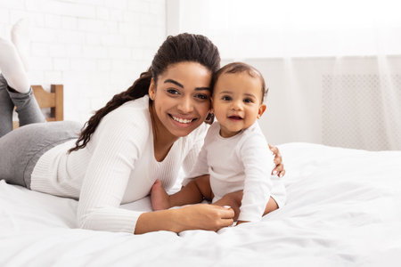 African Mother Hugging Baby Toddler Sitting On Bed Indoors