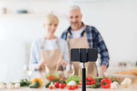 Smiling middle aged man and lady preparing salad and looking at smartphone webcam in kitchenの写真素材