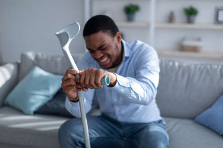 Young black man suffering from acute pain, leaning on crutch at home, selective focusの写真素材