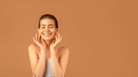 Charming young woman with nourishing cream on her face over brown studio background, banner design with free spaceの写真素材