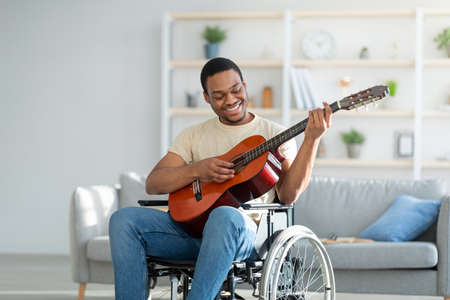 Handicapped black guy in wheelchair playing guitar and smiling at living room. Stay home activities
