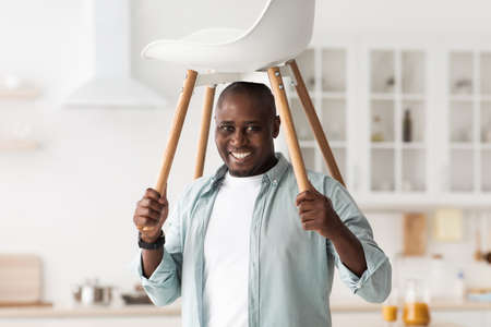 Furniture repair concept. Positive black man having fun with ready-made chair and raising it above his headの写真素材