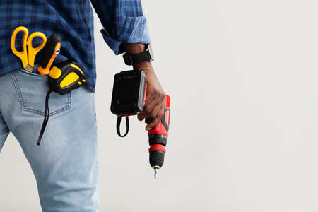 Black repairman holding electric drill in hand, in pocket measuring tape, screwdriver and scissors, copy spaceの写真素材